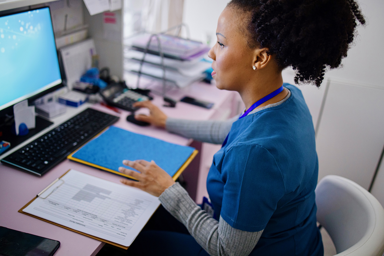 A woman in blue scrubs seated at a desk, using a computer while holding a clipboard with papers. A phone and office supplies are visible on the desk.