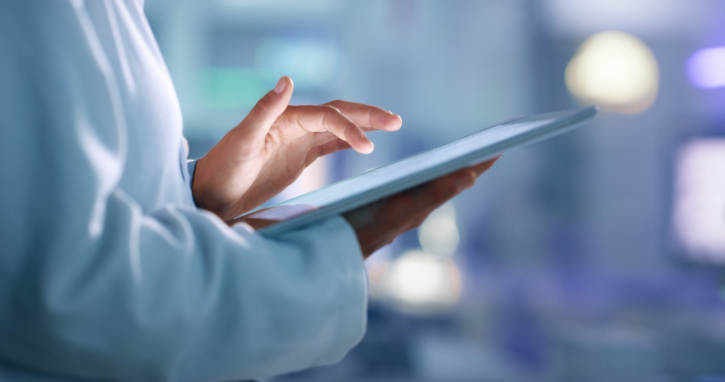 A close-up of a person's hand interacting with a touchscreen tablet, wearing a lab coat, in a softly lit modern workspace.