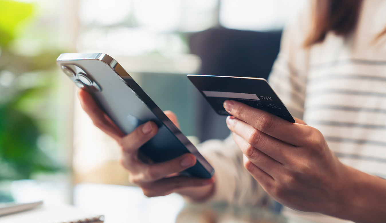 A person holding a smartphone in one hand and a credit card in the other, sitting at a table with a blurred background of indoor plants.