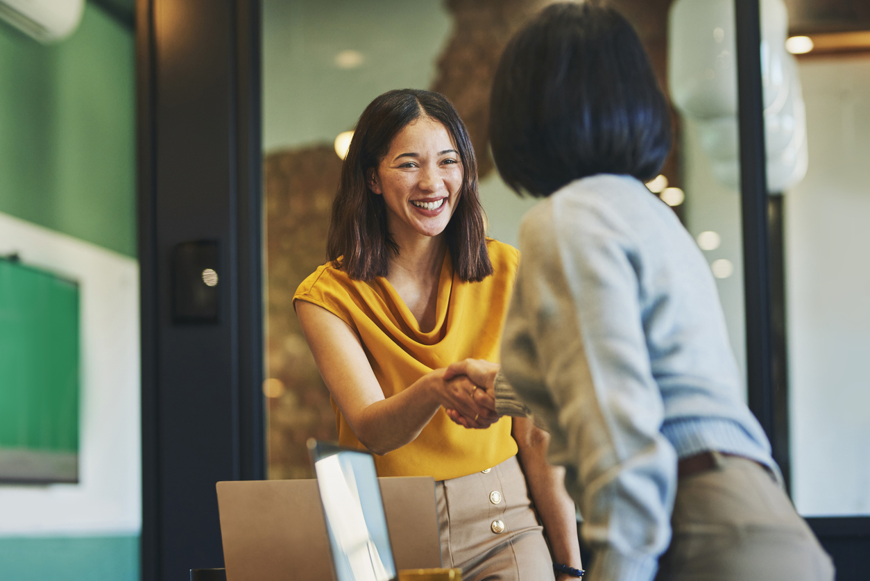 A woman wearing a yellow blouse is smiling and shaking hands with another woman in a gray sweater in a modern office setting.