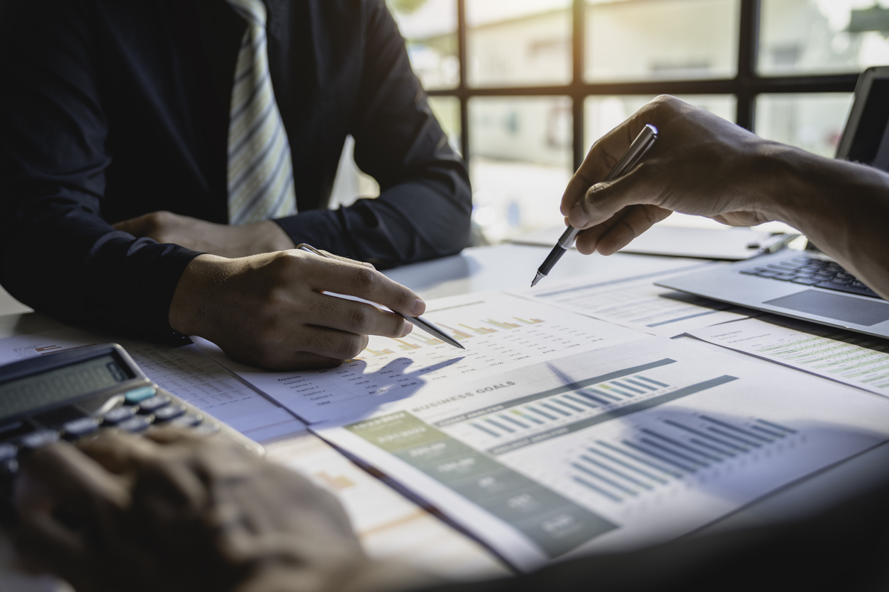 Two professionals reviewing financial documents and charts on a table, with one person pointing at the data and the other holding a pen, while a laptop is visible in the background.