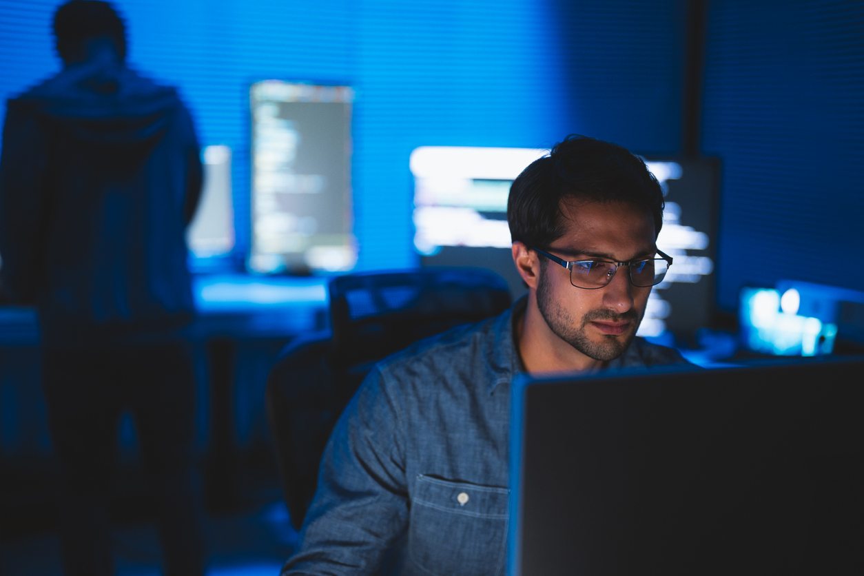 A man sitting at a desk, focused on his computer screen, wearing glasses. In the background, a person is seen standing near another computer, surrounded by a dimly lit environment with blue lighting.