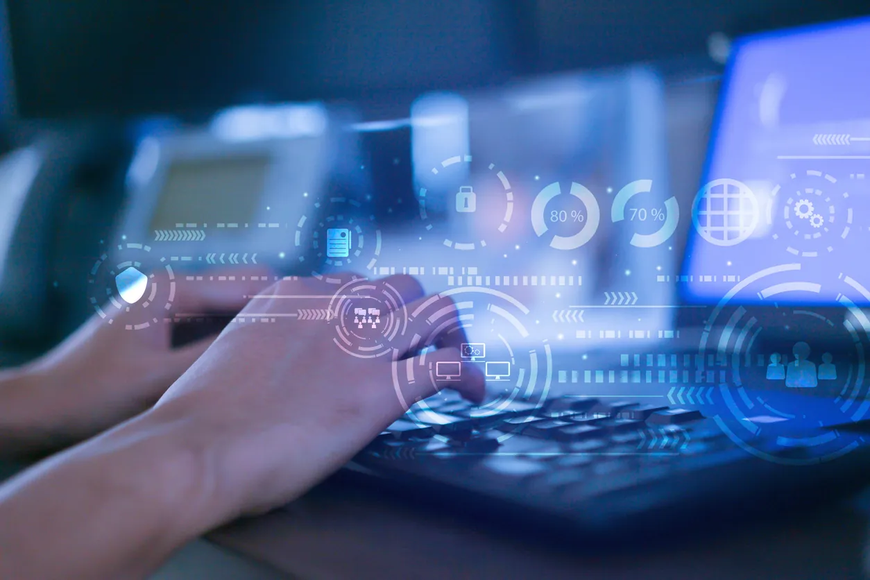 Close-up of a person's hands typing on a keyboard, with a digital overlay displaying various technology and data icons. A computer monitor is blurred in the background.