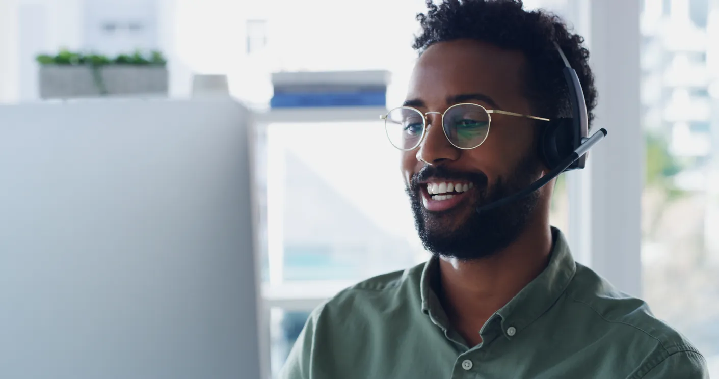 A man with curly hair and glasses wearing a headset, smiling while sitting at a desk in front of a computer screen in a bright, modern office.