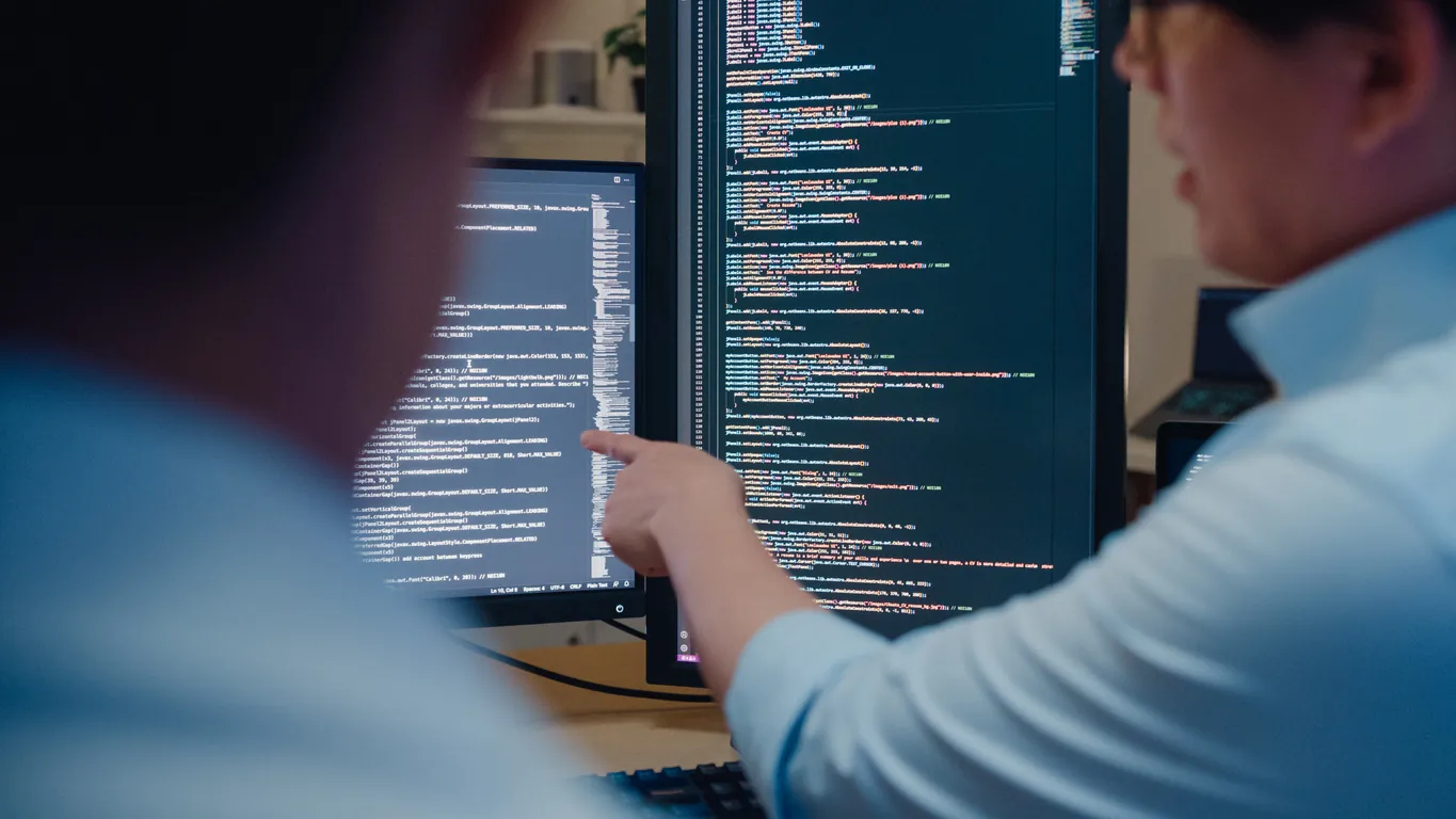 A person pointing at lines of code displayed on a computer screen, with another individual partially visible in the foreground. The setting appears to be a modern office space.