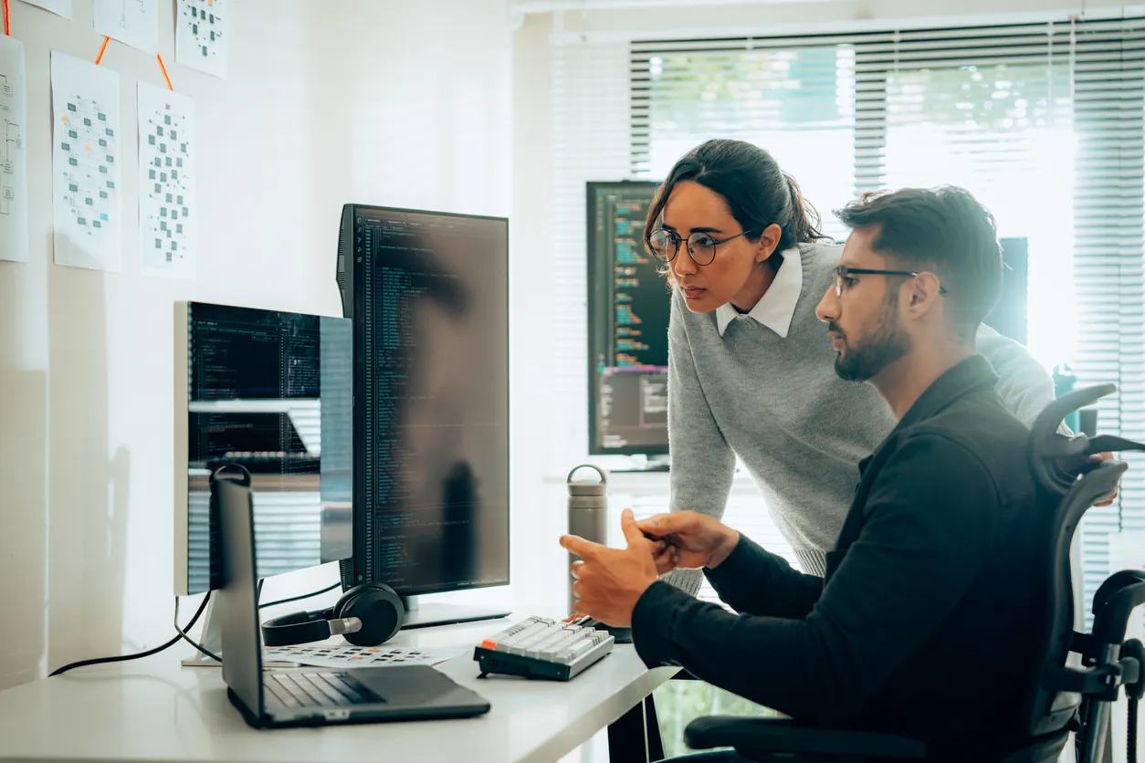 A woman and a man sitting in front of computer screens in a modern office, discussing coding on one screen while the other displays graphs. The man uses a wheelchair.