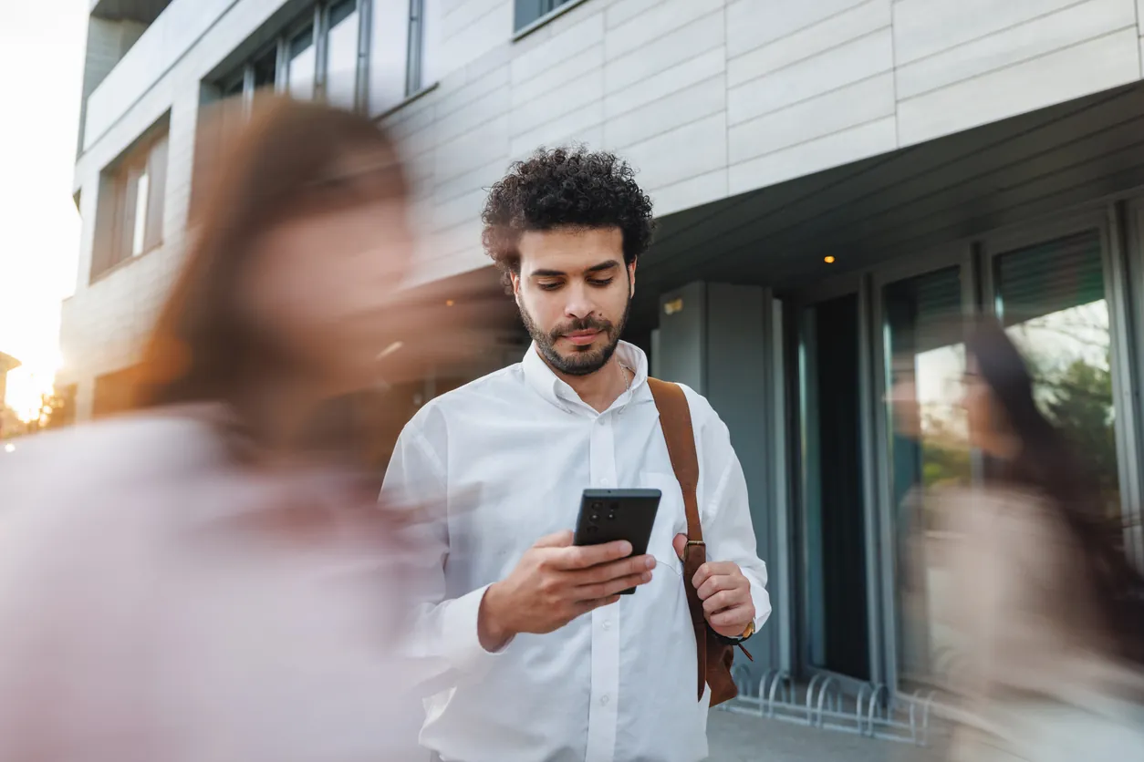 A man in a white shirt holding a smartphone while standing outside a modern building. Blurred figures of people are seen walking quickly around him.