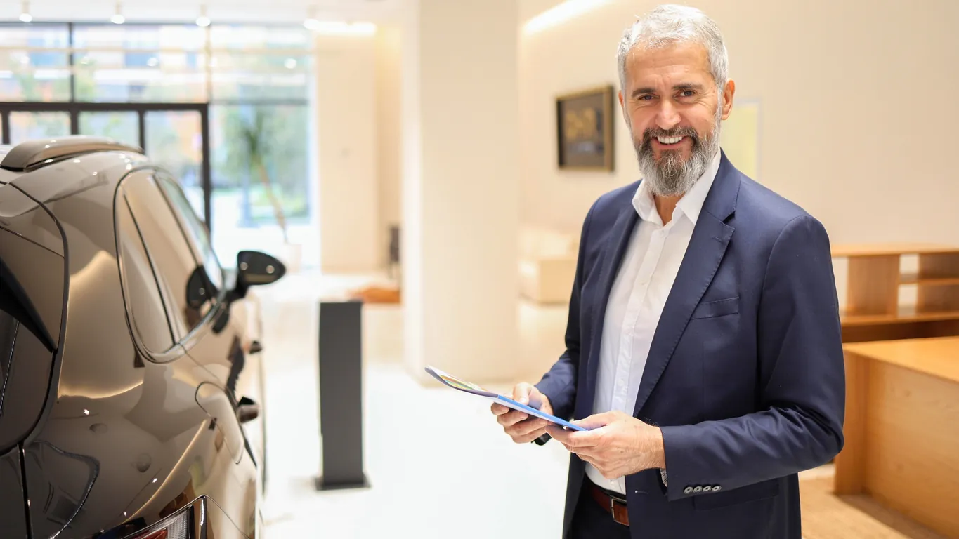 A middle-aged man in a suit stands next to a luxury car in a modern showroom, holding a document and smiling at the camera.