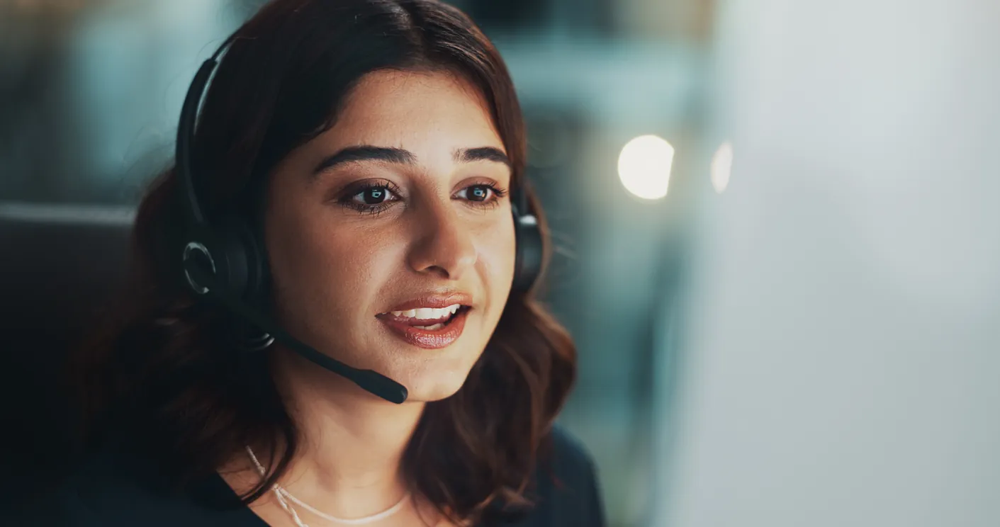 A woman wearing a headset engaged in a conversation at a desk, with a blurred background of an office environment.
