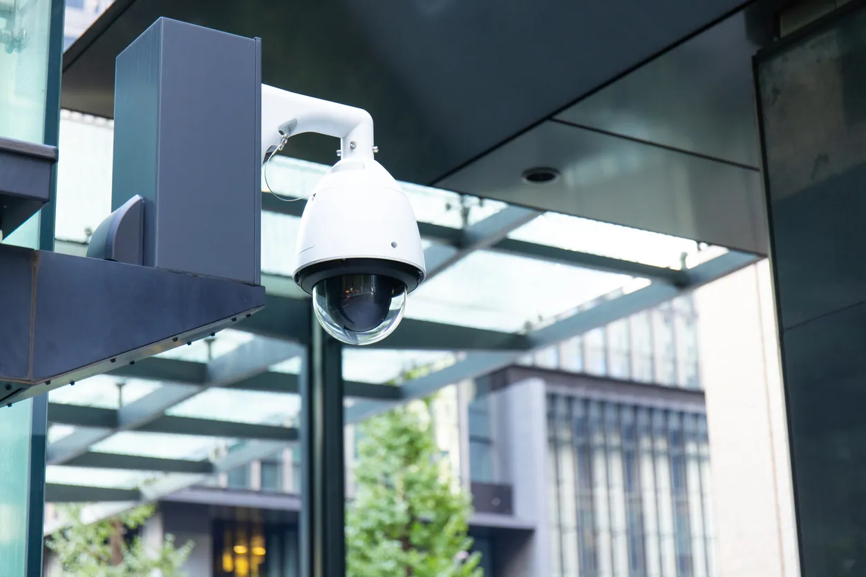 A close-up view of a white security camera mounted on a pole, surrounded by modern architecture and greenery in an urban setting.