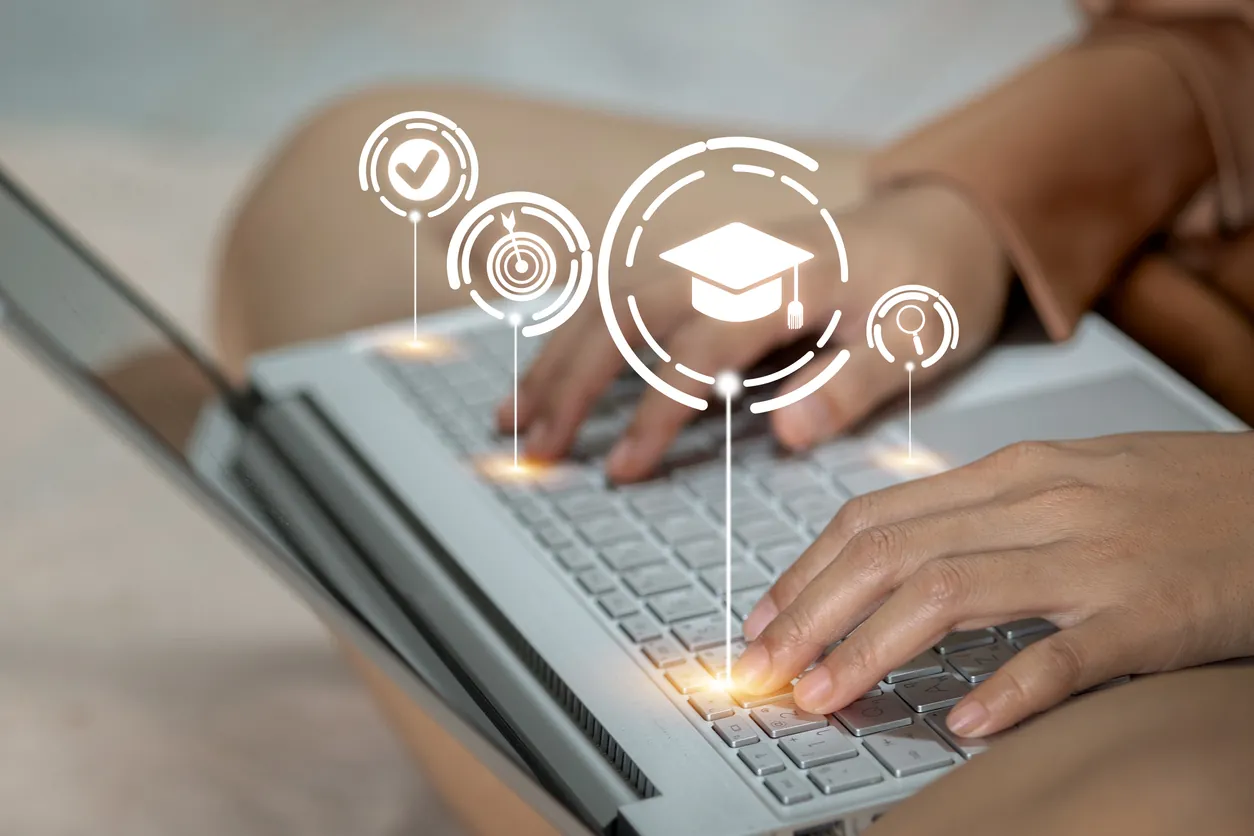 A person typing on a laptop keyboard, with graphic icons above representing education, achievement, and learning. The background is softly blurred, highlighting the focus on the hands and laptop.