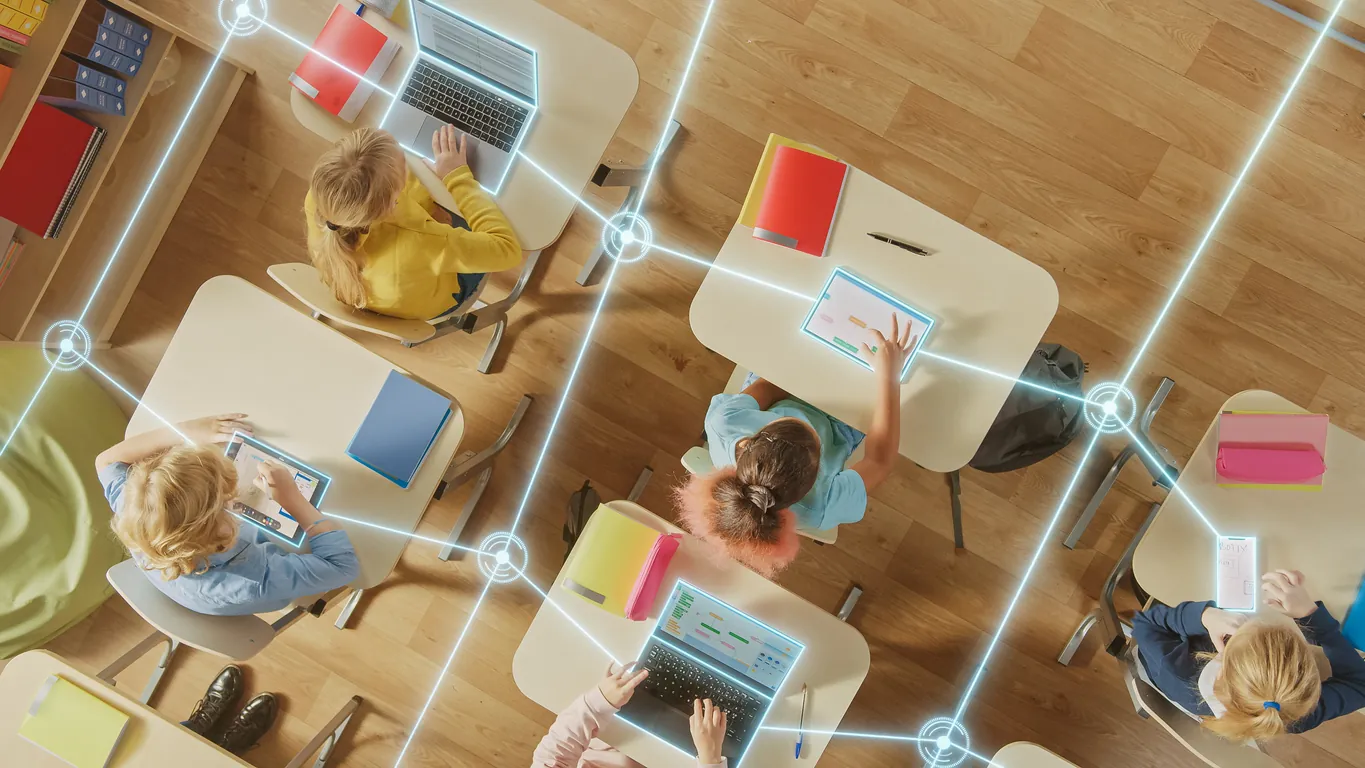 An overhead view of a classroom with students using laptops and tablets at their desks. Colorful folders and notebooks are visible on the desks, suggesting an engaged learning environment.