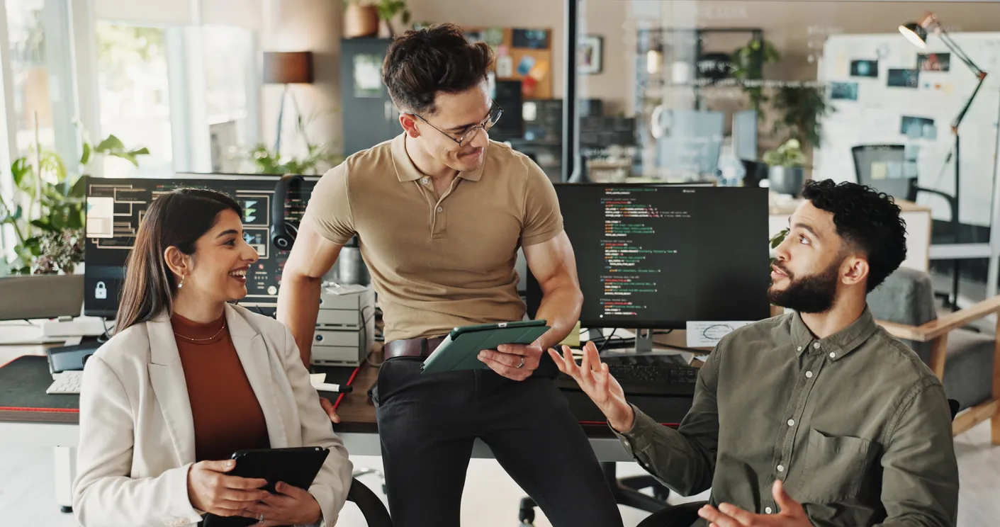 Three colleagues engaged in a discussion in a modern office. One man stands holding a tablet, while two others sit, one on a chair and one on the desk, each with a tablet in hand, surrounded by computers displaying code.
