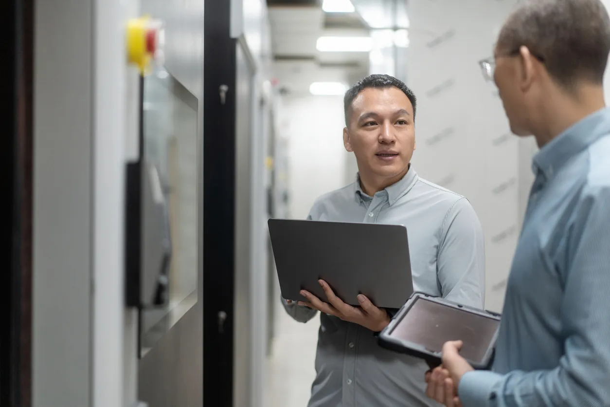 Two men discussing in a hallway, with one holding a laptop and the other holding a tablet. They appear engaged in a conversation, surrounded by blurred office partitions.