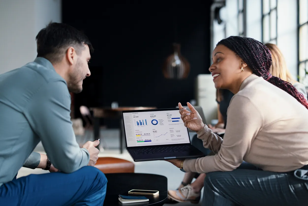 A man and a woman engaged in a discussion in a modern office space, with the woman pointing at a laptop screen displaying business analytics and charts.