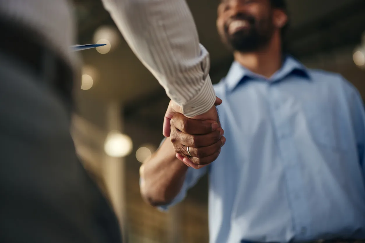 Two people shaking hands in a professional setting with a blurred background. One person is wearing a light blue shirt, while the other has a white long-sleeve shirt. Soft lighting enhances the friendly atmosphere.