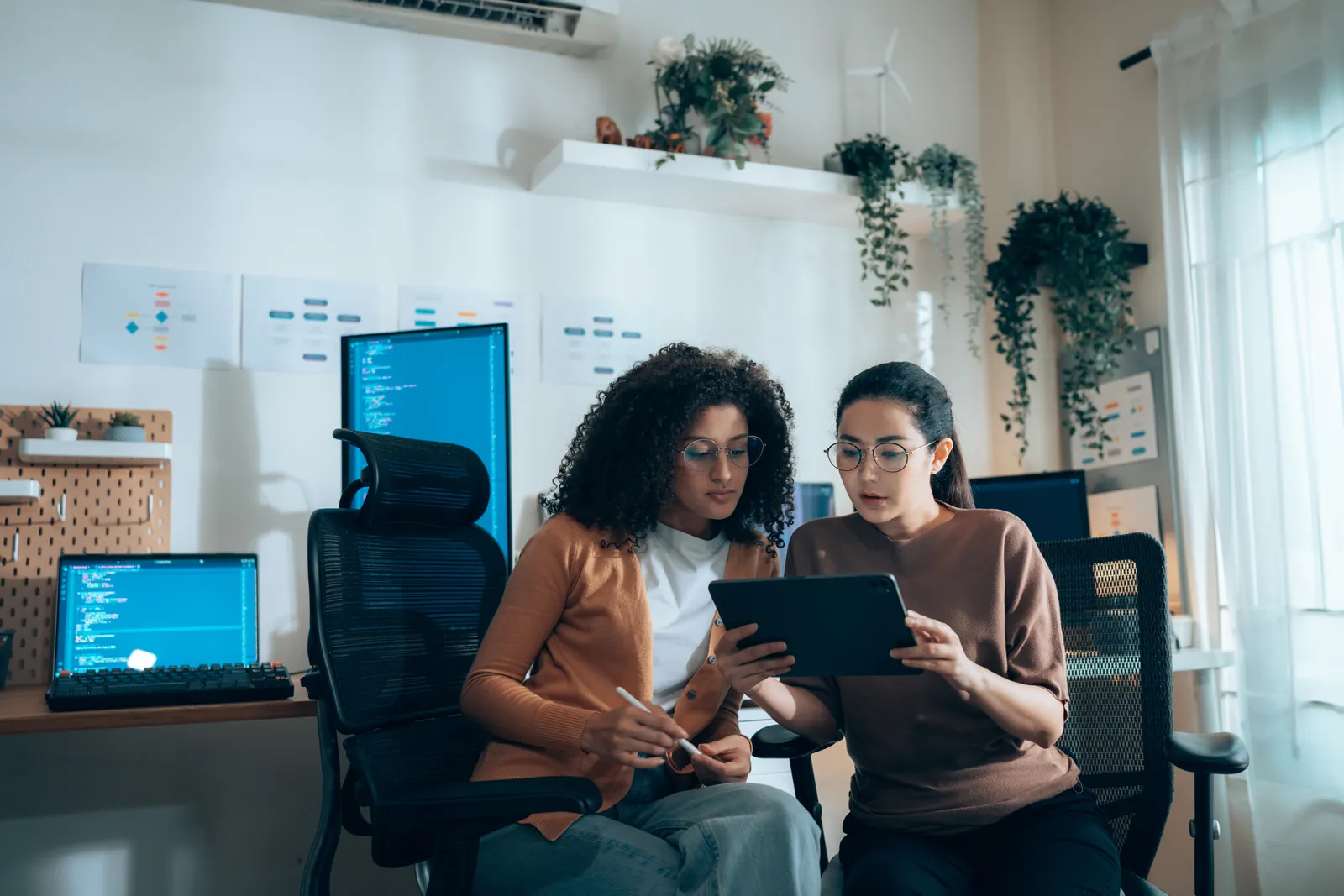 Two women collaborating at a desk in a modern workspace. One is holding a tablet while the other points at the screen. Laptops with code on screens are visible in the background, alongside plants and wall documents.