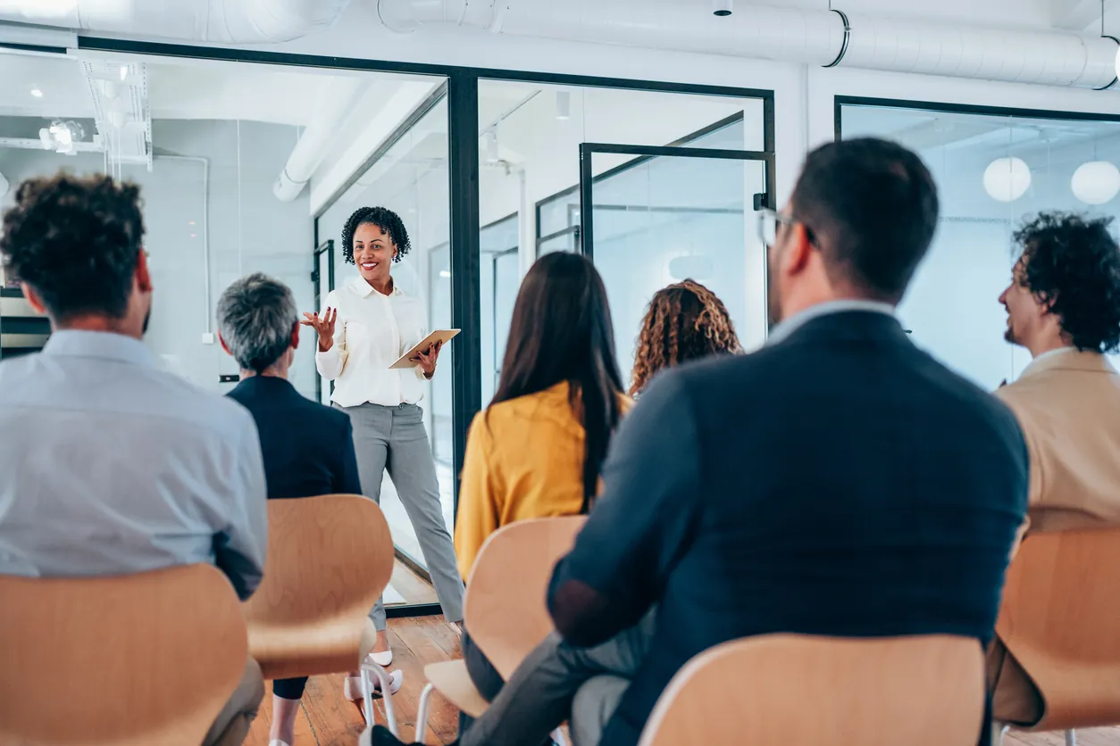 A woman standing and speaking in front of a group of seated colleagues in a modern office setting. The audience, diverse in appearance, is attentively listening.