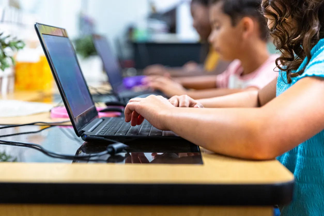 Children using laptops at a classroom table, focused on their screens. One child's hand is visible typing on the keyboard, while several other children are engaged with their devices in the background.