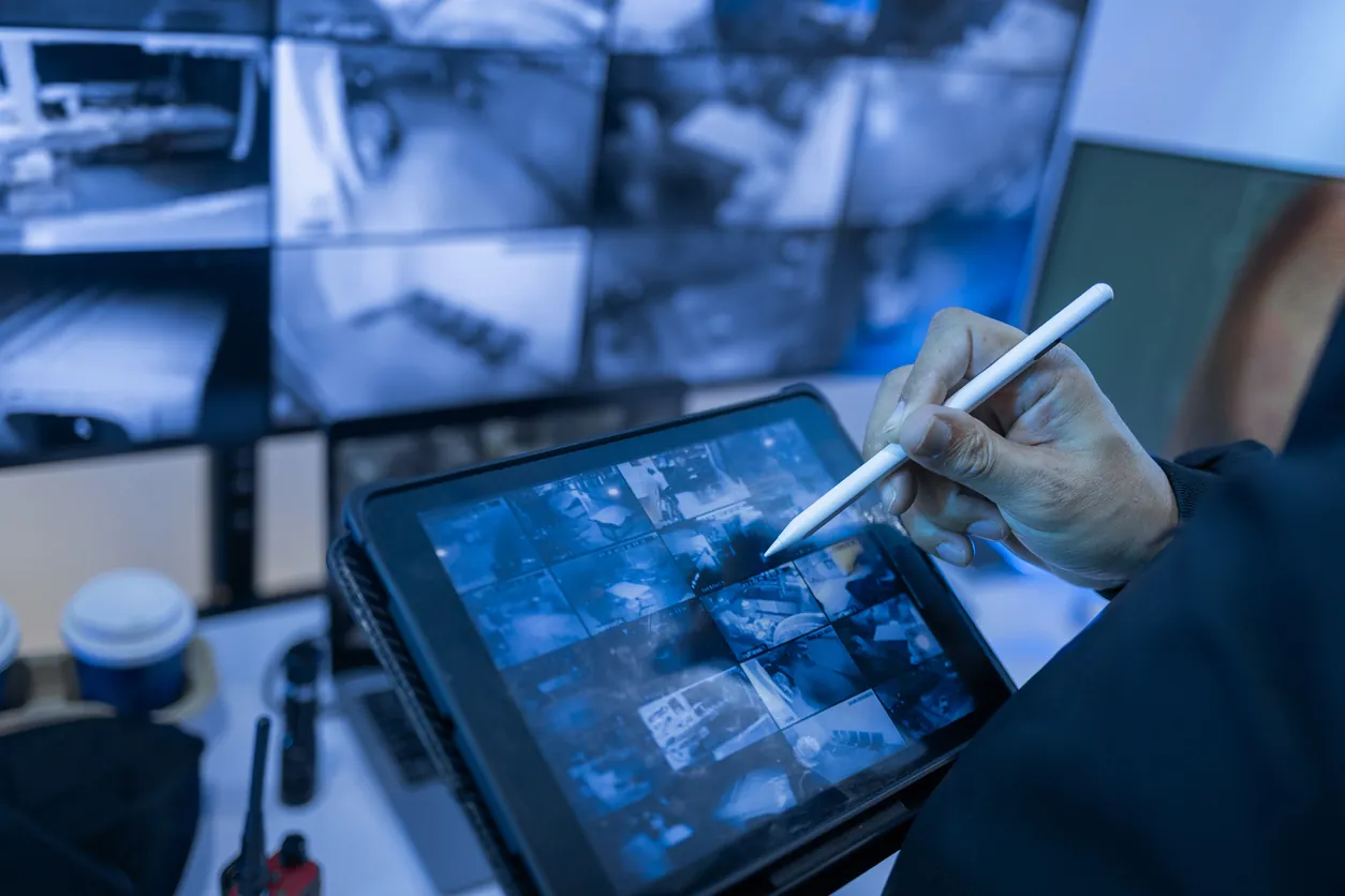 A person using a tablet to monitor security camera footage displayed on multiple screens in a control room. The tablet shows a grid of surveillance images.