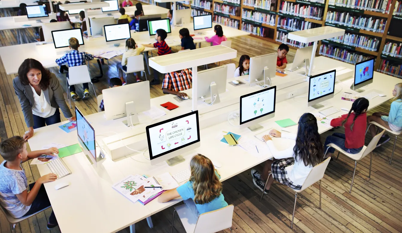 A classroom setting in a library with children using computers and engaged in various activities. A teacher is instructing a group at the front, while students work at individual stations, surrounded by bookshelves.
