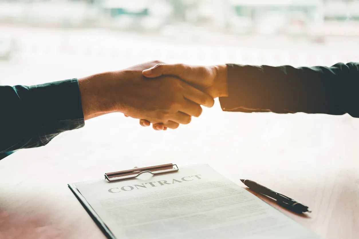 Two people shaking hands over a contract document on a wooden table, with a pen beside the document.